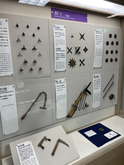 The image shows an exhibit of ninja weapons in the Iga Ninja Museum, located in Iga City, Japan. The display features various historical artifacts, including shuriken (throwing stars) of different designs, a higuruma-ken (incendiary dagger), a funso-tsubute (throwing weight), a kusari-gama (sickle with chain), and a fukiya-duto (blowpipe). The weapons are arranged in a clean, museum setting with informational text panels in Japanese and English describing each item. The overall environment is well-lit and organized, highlighting the historical significance of the ninja artifacts.