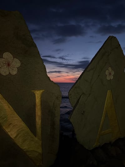 The image presents a close-up view of two stone structures adorned with golden symbols, set against a backdrop of a twilight sky and the ocean in Pererenan, Indonesia.

The stone structures feature a textured surface, with a flower motif and a golden "V" symbol on each. The upper stone is positioned at an angle, while the lower one is similarly oriented but contains the base part of the "V" in golden color.

The background reveals a dramatic sky transitioning from deep blue, indicating the transition between day and night. The ocean is visible in the narrow space between the stone structures, adding to the overall serene yet evocative mood of the photograph.