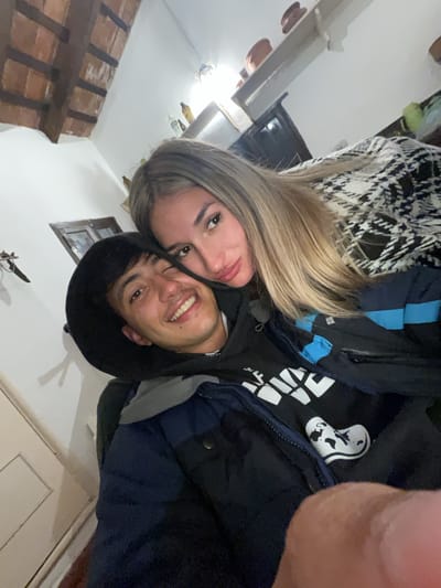 In El Rodeo, Argentina, a selfie captures a young couple embracing indoors. The woman, with blonde hair, leans her head on the man's shoulder, her face close to his. He smiles at the camera, his eyes squinting slightly. Both are dressed in casual, dark-colored outerwear. The background shows a rustic interior with exposed wooden beams on the ceiling and a white wall. Shelves with decorative items are visible on the wall, along with a framed picture and a portion of a doorway. The lighting suggests an indoor, possibly evening setting.