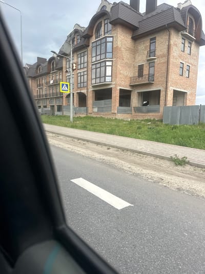 The image shows a view from inside a car, looking out onto a street in what appears to be Pidhorodnie, Ukraine. The primary subject is a large, multi-story brick building under construction, located to the left of the frame. The building features architectural details such as arches, balconies, and intricate roofing. A pedestrian crossing sign is visible near the building, indicating a crosswalk. To the right of the building is a strip of grass and a sidewalk, bordering an asphalt road. A white stripe marks a lane on the road. The car's interior is visible in the foreground. The sky is overcast. There are no people visible in the scene.