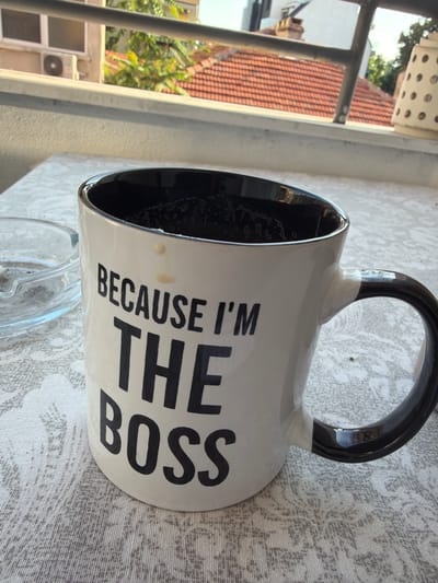 A white mug with black text that reads "BECAUSE I'M THE BOSS" sits on a table covered with a patterned cloth. Next to the mug is a glass ashtray. In the background, a balcony railing overlooks a street with buildings and a tiled roof. The lighting suggests it is daytime, possibly late afternoon.