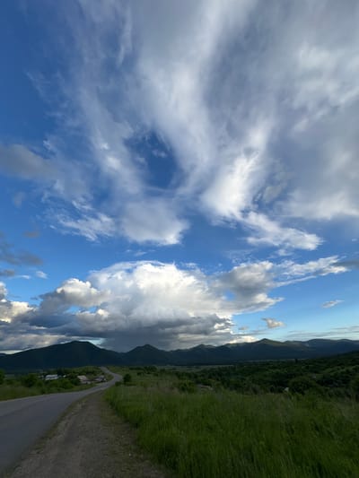 A view from a roadside in Теплоозёрск, Russia. The road curves away to the left, bordered by green grass on the right and a dirt shoulder on the left. In the distance, rolling green hills are covered in trees, with several dark, ominous clouds gathered above them. The sky above is a vibrant blue, filled with scattered white clouds, some wispy and others more substantial. A few small buildings with white roofs are visible nestled amongst the trees to the left of the road.