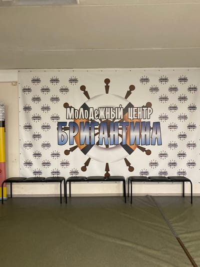 The image shows a banner for the "Brigantine Youth Center" located in Danilov, Russia. The banner features a white background with a repeating pattern of stylized images resembling gears or mechanical components. The center's logo consists of a ship's wheel shape with the Russian text "Молодёжный Центр Бригантина" written in bold, blue letters within a white circle. A row of black metal chairs is positioned beside the banner. The surroundings appear to be an indoor space with white walls and a dark-colored floor.