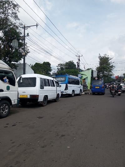 The image displays an urban street scene in Khajuripada, India, under an overcast sky. The asphalt road surface, appearing dark grey, exhibits several discolored patches. On the left side of the frame, the front left corner of a white truck is partially visible, featuring a blue character decal on its door. Adjacent to this truck, a white minivan with tinted windows and a yellow license plate bearing the alphanumeric sequence "F 2973 HA" is parked. Immediately behind the minivan, another white small bus, distinguished by a blue roof section and text that appears to read "PUTRA JAYA" above its windshield, is also parked.
Overhead, a complex network of black electrical wires spans the scene, supported by various utility poles. A decorative black street lamp post, featuring an ornate design and two light fixtures, stands on the left. Green foliage from numerous trees is visible behind the parked vehicles and structures along both sides of the road. In the mid-ground on the right, a light green building with a partial blue facade or roof structure is present, alongside a blue and white sign.
Further down the road on the right, a blue compact car is parked. Active traffic is observed in the mid-to-far right, consisting primarily of motorcycles. At least two motorcycles are moving towards the camera; the lead rider is wearing a white helmet. Additional vehicles, including a silver SUV, are visible in the background traffic flow. No direct interactions between individuals are discernible, and no pedestrians are clearly visible.