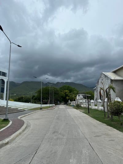 This image depicts a street scene in Oslob, Philippines, with a dramatic, overcast sky dominating the upper portion of the frame. The sky is filled with heavy, dark clouds, suggesting an impending storm or recent rainfall. In the foreground, a paved road stretches into the distance, bordered by a grassy verge and a white, ornamental fence. To the left of the road, there is a building with a white facade and dark windows, featuring a set of outdoor stairs. Further down the road on the right, a white stone church with a pitched roof is visible. Lush green mountains form the backdrop, partially obscured by the low-hanging clouds. Several streetlights are strategically placed along the road. The overall atmosphere is somber due to the weather, yet the architecture and natural landscape suggest a tranquil, possibly historical, location.