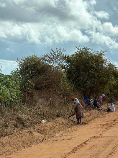 The image shows a group of people in a rural setting, likely in Pemba, Mozambique. Several individuals are resting or sitting on the ground near a dirt path. One person is standing and using a tool, possibly for digging or cultivating the land. The environment includes trees, vegetation, and a cloudy sky. The ground is dry and appears to be sandy or earthen. There is a white object discarded on the ground, possibly a piece of trash.