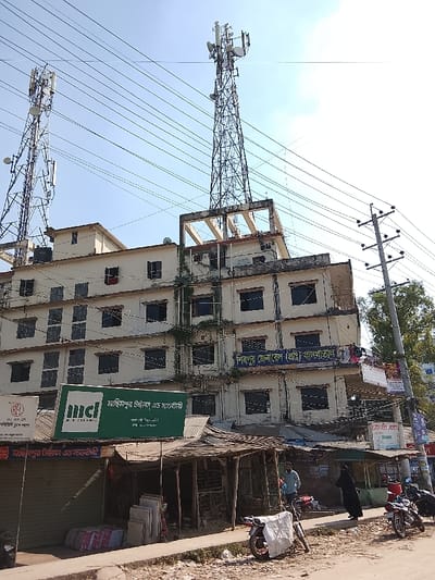 A five-story building is topped with a skeletal communication tower. Another communication tower stands to the left. Power lines crisscross the sky. Below, signs indicate shops and a private hospital, "Shipur General Hospital (Pvt.)". Shops with corrugated roofs and motorbikes line the street. An advertisement for "Machimpur Tiles and Sanitary" is visible on a green sign. Another sign references "RFL MIR CERAMIC" and "Machimpur Tiles".