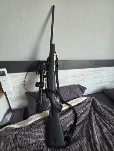 The image shows a rifle with a scope and sling leaning against a wall, likely in a bedroom. The rifle is black and appears to be a bolt-action rifle. The scope is mounted on top of the rifle. The sling is made of black leather and is attached to the rifle. The wall behind the rifle is white and textured. There is a headboard with a gray and white patterned design behind the rifle and a bed with pillows and a blanket. A remote control is visible on a shelf to the left of the rifle. The scene is indoors and does not provide any specific information about the city of Suvorovo, Bulgaria, or any people.