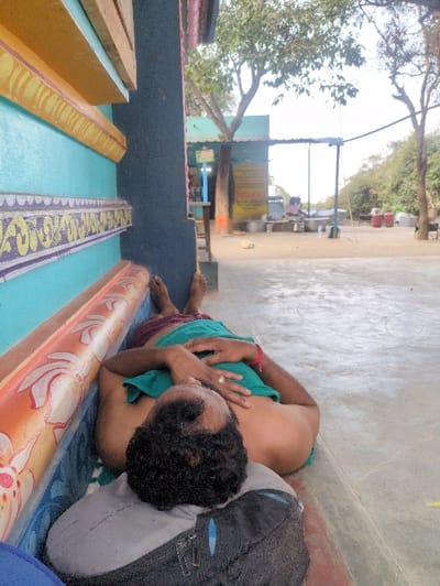 A man is depicted lying on his back on a light-colored concrete surface, using a grey and black backpack as a pillow. He has dark, curly hair and wears a maroon lungi and a teal-colored cloth covering his torso, with a silver ring on his left hand. His bare feet are visible. He is positioned alongside a brightly painted wall featuring blue, orange, and yellow sections with decorative patterns, adjacent to a dark blue pillar. The scene is outdoors in Kadaladi, India. In the mid-ground, a small structure with a light blue roof and signage is visible under trees. A light-colored dog lies on the ground in front of this structure. Further back, beneath a makeshift canopy, a person is seated near several large metallic vessels. Red gas cylinders are visible on the right side of the background, within an open area containing sparse trees against a bright sky.