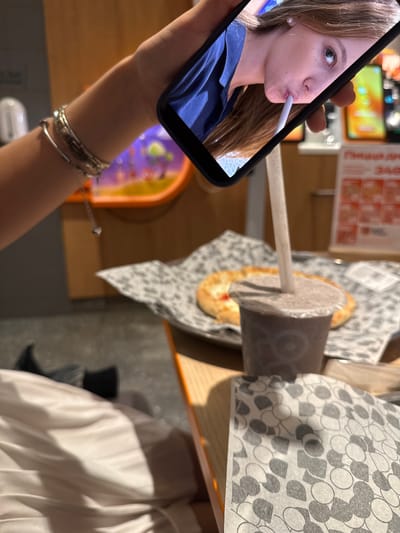 A person's arm, adorned with bracelets, holds a smartphone displaying a selfie of a young woman with long brown hair. The woman is looking at the camera and holding a white straw to her mouth. In the foreground, a cup with a straw and a plate with a flatbread topped with cheese and a red sauce are visible on a table. The background suggests an indoor setting, possibly a fast-food establishment, with blurry images of colorful displays and signage that may be in Russian. The overall scene captures a moment of casual dining and social media interaction. The location is Chusovoy, Russia.