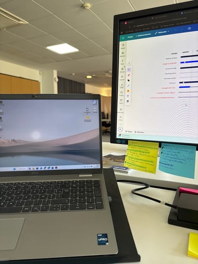 This image depicts a professional office workspace in Lièpvre, France, viewed from a slightly elevated perspective. In the foreground, a silver Dell laptop featuring an "Intel vPro" sticker rests on a black cooling stand. Its screen displays a stylized landscape wallpaper with a pale sun over water, and the Windows 11 taskbar shows the time "14:07" and date "20/02/2023." To the right, a black external monitor is active, showing the Canva.com interface with a project management timeline for "JANVIER" (January), listing tasks in French such as "Réseaux sociaux," "Google et social ads," "Articles de blog," and "Shooting photos." Two handwritten sticky notes are affixed to the lower-right bezel of the external monitor: a yellow one on the left contains "Infos:" and bulleted notes in French, while a blue one on the right contains "Échanges" and additional French notes, including "Inde." A black cable connects a peripheral to a black power strip or hub on the white desk surface. The background reveals a standard office environment with white acoustic ceiling tiles, recessed square LED light panels, and a visible circular smoke detector. Distant office furniture and partial reflections are discernible, indicating a larger open-plan office setting. No individuals are present in the frame.