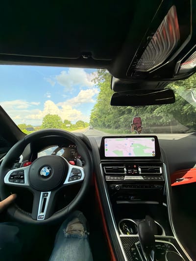 The image captures a first-person perspective from the driver's seat of a modern BMW M-series vehicle, situated on a road in the vicinity of Baiachevo, Bulgaria. The driver's left hand is visible on the black leather steering wheel, which features a BMW emblem, "M" badging, red M-mode buttons, paddle shifters, and integrated control buttons. The digital instrument cluster displays a current speed of 72 km/h, a total odometer reading of 23187 km, and an average speed of 44.8 km/h. Centrally located, an infotainment screen presents a navigation map with Cyrillic text, consistent with the specified Bulgarian location, and shows the time as 17:14. Below this screen, the climate control panels indicate a temperature setting of 22.0 degrees Celsius for both zones. The center console exhibits carbon fiber trim, a gear selector, an iDrive controller, and cup holders containing keys. A distinctive air freshener, shaped like a red-faced cartoon character talking on a phone, hangs from the rear-view mirror. The driver's left leg, clad in distressed blue denim jeans, is visible in the lower left. Outside the vehicle, a paved, two-lane road extends into the distance, bordered on both sides by dense green trees under a bright blue sky with prominent white cumulus clouds. Another vehicle is discernible further down the road. Distant hills are visible on the left horizon.
