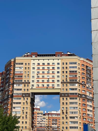This is a low-angle, vertical shot of a modern apartment building in Kotelniki, Russia, under a clear blue sky. The building, primarily constructed with tan and brown brickwork, features numerous windows with white frames. Some windows are rectangular and grouped in vertical columns, while others are smaller and square, arranged in horizontal rows. Many of the square windows have air conditioning units mounted below them.
The building's architecture is characterized by stepped levels and a central open section that reveals a patch of sky with some clouds. The topmost section of the building has a distinctive wavy roofline. In the lower right corner, the green leaves of a tree are visible, suggesting the presence of vegetation in the vicinity. The overall impression is of a bright, sunny day. No people are visible, and the image appears to capture a static view of the residential structure. There is no discernible text in the image.