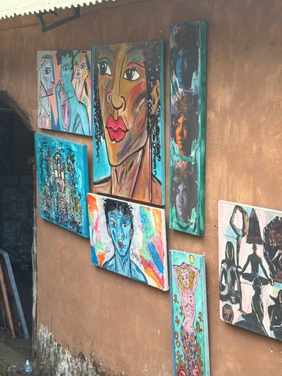 An outdoor wall in Tejakula, Indonesia, displays a collection of vibrant, colorful paintings. The wall itself is a textured, reddish-brown stucco, showing signs of age and some discoloration, particularly near the bottom where white and gray patches are visible. A corrugated metal overhang, supported by a dark metal bracket, runs along the top of the wall.
The paintings are arranged in a staggered, eclectic manner. On the left, a horizontal canvas features three stylized portraits of people with distinct facial features and bold outlines in shades of blue, pink, and yellow. To the right of this, a large, vertical painting prominently displays a close-up of a person's face with striking eyes, red lips, and dark braided hair. This painting is on a teal background.
Below the large face painting, a square canvas depicts a detailed, fantasy-like scene with a central white structure adorned with floral elements, all rendered in various shades of blue and green. Adjacent to this, another horizontal painting features a portrait of a person with blue skin and dark hair, set against a backdrop of abstract colorful lines and shapes.
To the right of the blue-skinned portrait, a tall, slender vertical painting showcases two stacked portraits of people with dark hair, against a green background. Below this, a smaller vertical painting displays a pink mermaid-like figure with blonde hair, against a turquoise background.
Further to the right, a larger, horizontal painting presents a collage of silhouettes of people in various yoga poses, with some text visible, including "Standing Poses" and "Salute." This painting has a pale pink background.
At the bottom left of the frame, leaning against the wall, are several unframed canvases or boards. A blue plastic water bottle with the brand "AQUA" is placed on the ground near the wall. The overall impression is of an art display in a rustic, possibly artistic, setting.