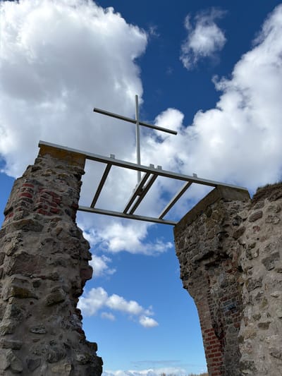 The image shows a low-angle shot of the ruins of the Salaspils church in Latvia. A stone structure with missing sections is visible, with a ladder-like framework and a cross protruding from the building. The sky is partly cloudy, with patches of blue visible behind the structure. The environment around the ruins is unclear due to the angle of the shot, but the scene captures the architectural remnants against a dynamic sky.