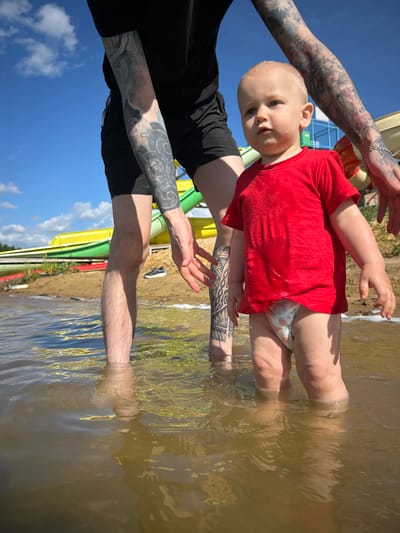 The image depicts a toddler standing in shallow, murky water. The toddler is wearing a red t-shirt and a diaper with a floral pattern. Behind the toddler, the legs of an adult are visible, one leg with extensive tattoos. The adult's hand is reaching towards the toddler. In the background, there are colorful water slides, a sandy shore, and a blue sky with white clouds. The location is identified as the city of Yubileynyy, Russia. The overall scene suggests a recreational setting, possibly a water park or a lakeside area on a sunny day.