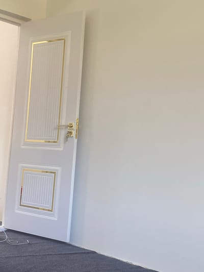 A white interior door with gold trim is partially open, revealing a glimpse of a bright white room. The door features two rectangular panels, each with a ribbed texture and outlined by a thin gold border. A silver-colored door handle and lock mechanism are visible on the door, with two keys hanging from the lock. The door is set against a plain, light-colored wall. The floor in front of the door is covered with dark gray carpet. A white cable is lying on the carpet near the bottom left corner of the image. The scene appears to be located in a newly constructed or renovated building.