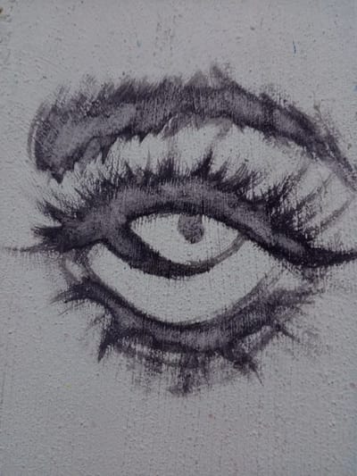The image is a close-up of an eye painted in charcoal on a textured, likely concrete, wall. The eye is stylized with thick, expressive eyebrows and eyelashes, and a visible iris. The artwork appears to be part of a larger mural or graffiti piece, given the texture of the wall, but only the eye is visible within the frame. The location, Parroquia Ocumare, Venezuela, is inferred, and there are no observable people or interactions in the scene.