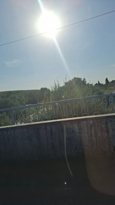 A bright sun is visible in the upper left quadrant of the image, casting a strong glare and a beam of light diagonally across the sky. A thin black wire stretches horizontally across the sky. The sky is clear and blue. Below the sky, a railing made of light-colored metal bars, some curved and some straight, runs horizontally across the middle of the image. Behind the railing, a body of water is visible, with some vegetation growing along its banks. The landscape behind the water consists of rolling hills covered in trees and shrubs. In the foreground, a concrete barrier is visible, running horizontally across the bottom of the image. The lower portion of the image is dark, with a small, bright green circular object visible towards the bottom right. The scene appears to be outdoors in a natural setting.