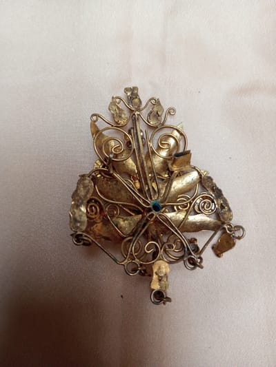A single metallic object is depicted against a light beige, subtly textured fabric surface, located in Qarabucaq qəsəbə, Azerbaijan. The object is an irregularly shaped piece of ornate craftsmanship, composed of aged, gold-toned metallic material. Its design features intricate wirework, including scroll motifs and interconnected elements, alongside flattened leaf-like components. A small, round, blue-green cabochon-like element is centrally embedded. The metallic surface exhibits varying degrees of tarnish and discoloration. Structural inconsistencies, including areas of deformation and partially detached or missing components, are visible along the object's periphery. No people or other active subjects are present.
