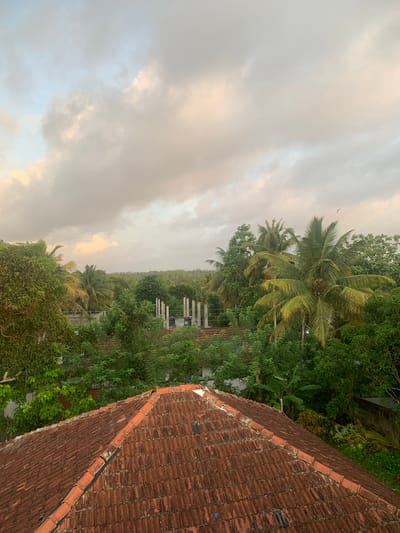 The image depicts an aerial view of a tiled roof in Karandugoda, Sri Lanka, with a lush, dense green landscape stretching into the distance. The foreground is dominated by the terracotta tiles of the roof, which show signs of age and weathering. Beyond the roof, a variety of trees, including palm trees and mango trees, create a vibrant green canopy. In the middle ground, construction is underway with concrete pillars and water tanks visible. The sky above is filled with a mix of light and dark clouds, with a hint of soft, diffused sunlight filtering through. There are no people or animals visible in the scene. The overall impression is one of a tropical, somewhat undeveloped, rural or suburban setting.