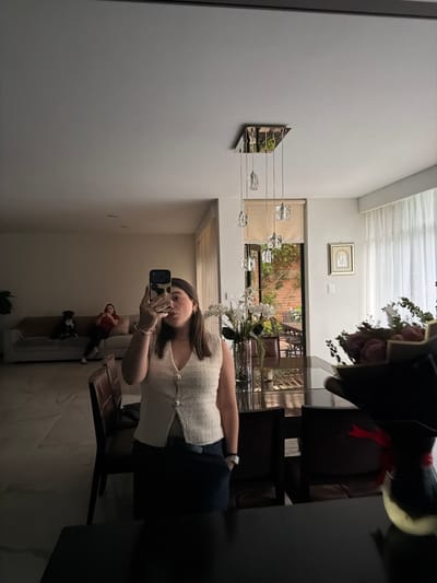 A young woman is taking a selfie in a mirror. The woman is wearing a white shirt and dark pants. The mirror is in a room with a dining table. There is a bouquet of flowers on the table. The room is in a building in Barrio de Nuevo León, Mexico. There is a modern style pendant light on the wall. There is a picture hanging on the wall above the doorway. A small dog is resting on a chair in the background.