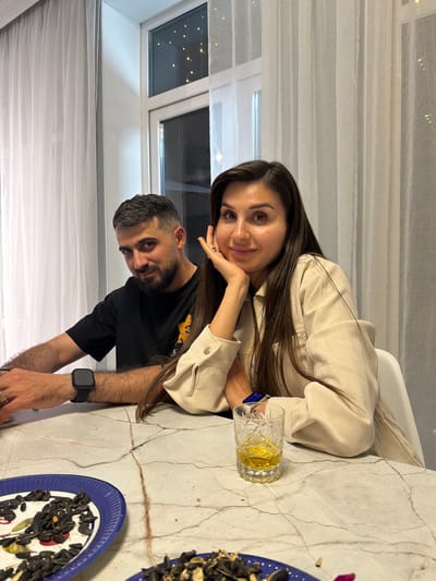 The image depicts a woman and a man at a table. The woman, with long dark hair, rests her head on her hand. She wears a light-colored jacket. Beside her is a glass containing a yellow liquid. The man, with short dark hair and a beard, wears a black t-shirt and a smartwatch. Two plates of what appear to be sunflower seeds are on the table. The background shows a window with a curtain. The location is Khoroshovskiy, Russia.
