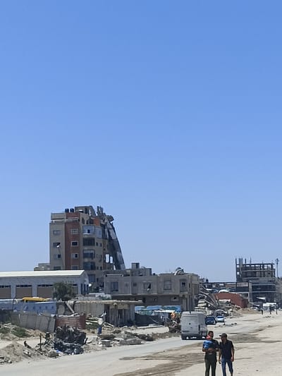 An aerial view of damaged buildings in Gaza City, Gaza Strip, Palestine, depicts a scene of urban devastation under a clear blue sky. In the foreground, a dirt road is visible, with debris scattered along its edges, suggesting recent destruction. Two men are walking on the road, one carrying a baby. Behind them, a white van is parked. Further down the road, several cars are seen.
The middle ground is dominated by a severely damaged multi-story building, its facade partially collapsed, revealing exposed interior structures. Adjacent to it are other buildings, some appearing intact and others showing signs of damage. A long, tilted structure, possibly a ramp or a section of a collapsed building, extends from the damaged building downwards. In the distance, a partially constructed building with visible scaffolding stands as a stark reminder of ongoing development amidst the destruction. The overall environment appears arid and dusty, consistent with the Gaza Strip's landscape.