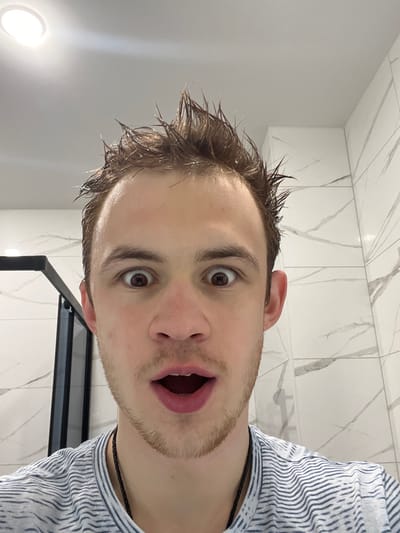 A close-up portrait captures a light-skinned adult male, approximately 20-30 years old, indoors within a bathroom setting in Tliustenkhabl', Russia. The subject's brown hair is wet and spiked upwards. His facial expression indicates extreme surprise or shock, characterized by wide-open brown eyes with dilated pupils, raised eyebrows, and an open mouth. Light stubble is visible on his chin and jawline. He wears a light-colored t-shirt with thin, wavy horizontal stripes and a black cord necklace. The background consists of white and grey marble-patterned wall tiles. A recessed circular ceiling light fixture is visible in the upper left, and a dark, possibly black, structural frame, likely part of a shower enclosure, is partially present on the left side of the frame.