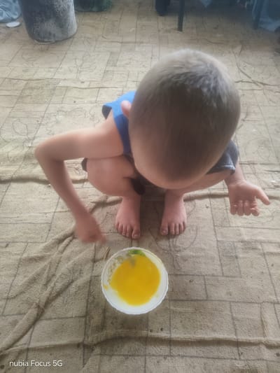 In Sestrynivka, Ukraine, a young child is crouched on a patterned mat, facing a bowl of yellow liquid. The child wears a blue top and dark shorts, with their head down as they observe the contents of the bowl. Their bare feet are visible, one resting on the mat and the other slightly raised. An arm is extended towards the bowl, and a hand is submerged in the liquid. The background features a cylindrical object and the floor is covered by a textured mat with a grid pattern. The lighting suggests an indoor or shaded outdoor environment.