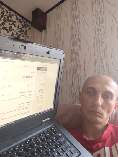 The image shows a man in his late 30s or early 40s with a shaved head, looking directly at the camera. He is wearing a red t-shirt and has a thin build. He is seated in front of an open laptop, which is angled towards him. The laptop screen displays a webpage with text in what appears to be Russian.
The setting appears to be indoors, possibly a home environment. The wallpaper behind the man has a vertical striped pattern. In the upper left corner of the image, a dark, rectangular object is mounted on the wall, possibly a speaker or a ventilation unit.
The man's expression is neutral, perhaps slightly tired or pensive. The lighting suggests it could be daytime, but the specific time is not clear. The overall atmosphere is casual and domestic. The visible text on the laptop screen includes what seems to be financial information or account details, with Cyrillic characters. Phrases like "ДЕНЬГИ" (money) and numbers with currency symbols are discernible.
The image seems to be a selfie, taken by the man himself. The focus is on the man and the laptop, with the background providing context. There are no strong indicators of time of day or weather conditions. The location cue "Bor, Russia" is provided.