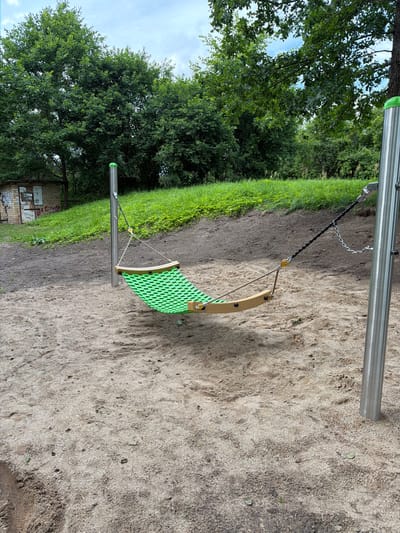 The image depicts a playground scene in Tēraudi, Latvia. In the foreground, a sand-covered ground transitions into a grassy embankment. A green net hammock with wooden edges is suspended between two metal poles, one on the left and one on the right, with ropes and chains connecting them. In the background, lush green trees and foliage dominate the landscape, with a small, weathered brick structure visible on the left side of the frame. The sky above is partly cloudy. There are no people or animals visible in the image.