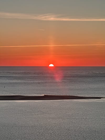This image captures a serene sunset over the ocean in La Teste-de-Buch, France. The sun, a vibrant orange disc, is partially submerged below the horizon, casting a bright reflection on the water. The sky transitions from a deep orange near the horizon to a lighter peach and then a pale blue at the top, with thin, wispy clouds and two contrails streaking across it. The ocean itself is a deep gray, with gentle ripples on its surface. In the foreground, a dark, sandy landmass, likely a dune or a sandbar, separates two distinct bodies of water. The overall scene is peaceful and tranquil, bathed in the warm glow of the setting sun.