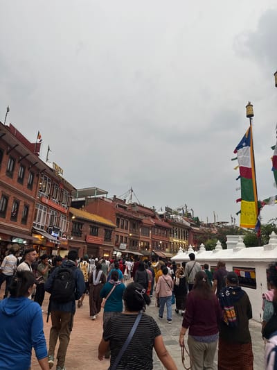The image shows a street scene in Gokarneshwar, Nepal. A crowd of people is present, some appearing to be walking or milling about. Buildings with brick facades line the street. A colorful flag is visible. The sky is overcast.