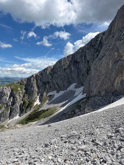 The image depicts a rugged mountain landscape under a partly cloudy blue sky. The foreground is dominated by a scree slope composed of numerous gray rocks of varying sizes. A diagonal band of snow stretches across the middle ground, interspersed with patches of green vegetation and small shrubs clinging to the rocky terrain.

In the background, a steep, imposing rock face rises dramatically. Its surface is textured with cracks and fissures, indicating its geological composition. Further in the distance, rolling green hills and mountains are visible under a sky filled with cumulus clouds. There are no people or man-made structures visible in the image. The scene conveys a sense of remote wilderness and the raw beauty of a mountainous environment. The specific location of Pašina Voda, Montenegro, is not visually identifiable from the image itself, as it lacks any identifying landmarks or signs.