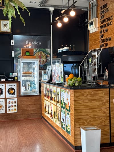 The image shows the interior of a cafe, likely in Tha Chana, Thailand, characterized by its warm wooden decor and a selection of beverages displayed. The scene is well-lit, with industrial-style pendant lights hanging from the ceiling, casting a warm glow. On the wall to the right, a menu board lists various coffee drinks like "COFFEE," "ESPRESSO," "LATTE," "CAPPUCCINO," "AMERICANO," "MOCHA," "DRIP," "COLD BREW," and "MATCHA."
In the center of the image, a refrigerated display case showcases food items, illuminated by internal LED lights. Adjacent to it, a counter displays a basket of fresh oranges and limes, a magazine rack, and various promotional posters for drinks. To the left, more posters, including "THE BRIM," are visible, along with a "TIP BOX." The floor is a polished wooden surface. The overall atmosphere is inviting and suggests a popular spot for coffee and refreshments.