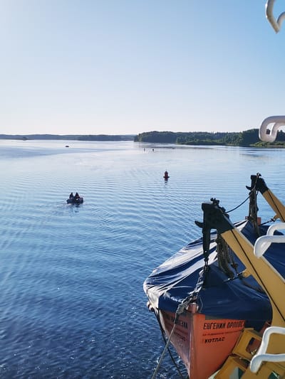 This image depicts a serene daytime scene on a wide, calm body of water in Ul'yanovo, Russia, under a clear, pale blue sky. The perspective is from a larger vessel, with its orange lifeboat, partially covered by a dark blue tarp, visible in the lower right foreground. The lifeboat is labeled "ЕВГЕНИЙ ОРЛОВСКІ" (Yevgeny Orlovsky), with "АО РЫБИНСКОЕ ПАРОХОДСТВО" (JSC Rybinsk Shipping Company) and "КОТЛАС" (Kotlas) also inscribed. Yellow davits and associated ropes secure the lifeboat, alongside stacked yellow crates.
In the mid-distance, two individuals are navigating the blue water in a small, grey inflatable boat, propelled by an outboard motor, generating a minor wake. Several red buoys or markers are distributed across the water, with one prominently visible to the right and another further to the left. The water surface exhibits gentle ripples extending from the various vessels. The background features distant shores densely lined with green forests of deciduous and coniferous trees, stretching across the horizon under the clear sky. A faint white contrail is discernible in the upper sky.