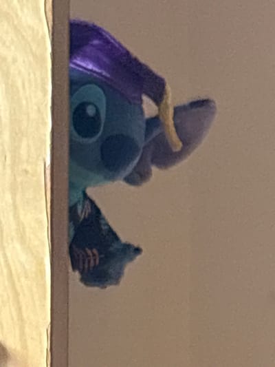 A blue and purple plush toy of the Disney character Stitch sits on a light-colored, possibly wooden, surface. The toy is positioned so that only its head, part of its ears, and part of its back are visible above the edge of the surface. The image is slightly blurry and has a muted color palette, giving it a soft and indistinct appearance. The background is a plain, light tan color.