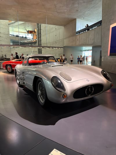 The image depicts the interior of the Mercedes-Benz Museum in Stuttgart, Germany. In the foreground, a silver 1955 Mercedes-Benz 300 SL Gullwing is parked on a dark grey floor. The car is facing towards the right of the frame, with its front grille and headlights clearly visible. To the left of the Gullwing, a red 1950s Mercedes-Benz 300 SL Roadster is partially visible. In the background, several people are seen walking around and observing the exhibits, including individuals on a raised walkway. Suspended from the ceiling is a vintage airplane. The museum's architecture is characterized by concrete walls and ceilings, with a modern and spacious design.