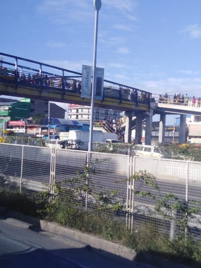 A pedestrian overpass is crowded with people on a sunny day. The bridge has yellow sides and black railings and features "Midea" advertisements. Below, a busy street has vans and other vehicles driving by, partially obscured by a wire fence with vegetation growing at its base. A tall lamppost stands beside the bridge. Buildings are visible in the background.