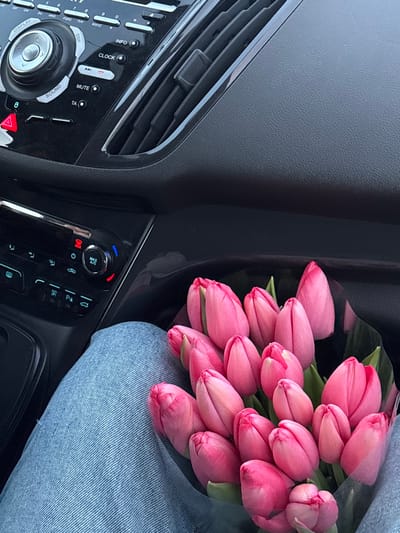 The image depicts the interior of a vehicle, viewed from a slightly elevated perspective. A person's lap, covered by light blue denim jeans, occupies the lower left and central portion of the frame. Resting on the lap is a dense bouquet of approximately 20-25 vibrant pink tulip buds, some with visible green foliage, wrapped in clear plastic or cellophane.
In the upper half of the image, the dark grey/black dashboard and central console of the vehicle are visible. The dashboard features a textured surface. Prominently displayed is a car stereo and climate control panel. The upper section of the console includes a main rotary knob, labeled "OK" in its center, surrounded by buttons such as "DISPLAY," "SOUND," "MUTE," "INFO," "CLOCK," "TA," and a red triangular hazard light button. Below this, a secondary control panel features additional rotary knobs, including one marked "MAX A/C," and various other climate and vehicle function buttons. Black air vents are visible to the right of the upper control panel.
The specific location within the city of Slavyansk-na-Kubani, Russia, is not discernible from the visual information presented in the image.