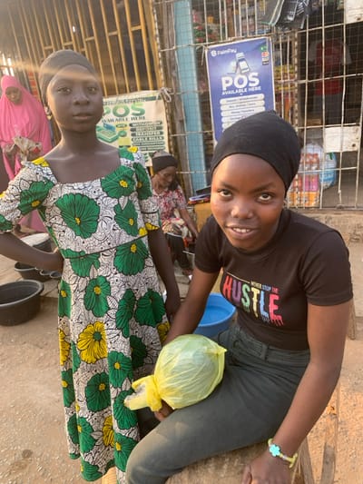 Two women pose for the camera in an outdoor market setting. One wears a black head covering and a black t-shirt with the words "NEVER STOP THE HUSTLE" printed on it. She is sitting and holding a plastic bag containing something round. The other woman stands beside her, wearing a colorful floral dress with a matching head covering. Market stalls and people are visible in the background, along with signs advertising POS services. The lighting suggests it is daytime.