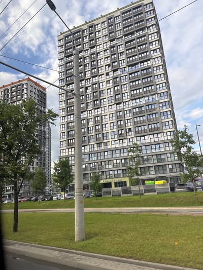 The image depicts a modern residential complex in Senitsky Sel'skiy Sovet, Belarus. Two tall, gray apartment buildings with repetitive window patterns dominate the scene. The building on the right is taller and wider, featuring balconies with dark railings. The building on the left is slightly shorter and has a distinct orange band near its roofline. A concrete utility pole stands prominently in the foreground, adorned with wires and a street lamp at its top. In front of the buildings, a well-maintained grassy slope leads down to a street. A wooden fence runs along the upper part of the slope, with a few trees scattered in front of it. Several cars are parked in the background on the street level. The sky is partly cloudy with patches of blue.