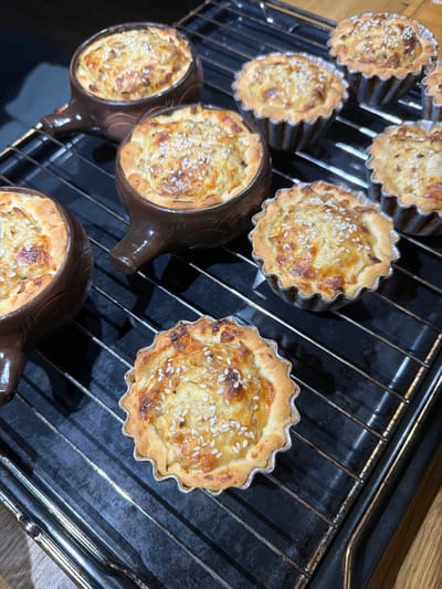 In this top-down, close-up shot, several savory tartlets are cooling on a black metal oven rack. The tartlets are nestled in fluted aluminum molds and some are also in decorative brown ceramic ramekins that resemble snails. Each tartlet features a golden-brown, flaky crust filled with a savory mixture. The filling appears to be a creamy base with visible pieces of what might be vegetables or meat, all topped with a generous sprinkle of sesame seeds. The tartlets have a slightly browned and bubbly appearance, suggesting they have recently been baked.
The setting is a kitchen, with the oven rack resting on what looks like a kitchen counter or the base of an oven. The lighting is warm and indoor, indicating the activity is taking place inside. The overall impression is one of freshly prepared food, likely a meal or appetizer. There are no people visible in the frame, and no visible text. The focus is entirely on the food and the cooking implements. The image captures a moment of domestic preparation and enjoyment of baked goods.