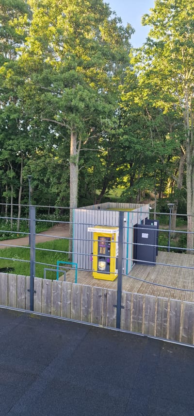 In the city of Vārzas, Latvia, a yellow vending machine is situated on a wooden platform surrounded by a fence. The vending machine displays the text "XXL KAFĪJA LIDZŅEMŠANAI" and "LOFFREDS". To the right of the vending machine, there is a dark-colored door, possibly for an enclosure. The scene is set against a backdrop of lush green trees, with sunlight filtering through the leaves. A black asphalt area is visible in the foreground, with a metal railing separating it from the platform and the vending machine. The sky is clear and blue, indicating a sunny day.