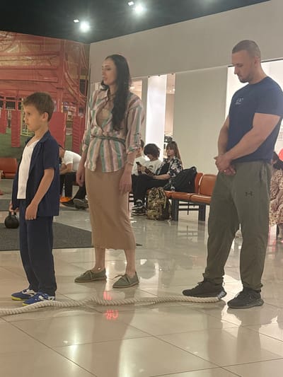 In the city of Tri Protoka, Russia, three individuals are positioned on a thick rope laid horizontally across a polished tiled floor. To the left, a young boy, dressed in a dark blue suit and white t-shirt, stands with his left hand holding a black kettlebell. Next to him, a woman in a vertically striped, multi-colored blouse and a tan skirt stands looking to her left. To the right, a man wearing a dark blue t-shirt with a white logo and olive-green pants stands facing away from the camera, with his hands clasped in front of him. In the background, several other people are seated on orange benches. One individual is looking at a phone, and another is wearing a patterned shirt. The environment appears to be an indoor public space with a large, orange-patterned mural visible on the wall behind the boy and woman. The lighting suggests an indoor, possibly commercial or recreational, setting.
