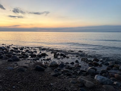 This media file depicts a serene coastal scene at Dronningmølle, Denmark, during twilight. The primary subjects are a rocky shoreline and the calm sea under a vast sky. The foreground is dominated by a collection of smooth, variously sized stones and pebbles scattered across the sand. Some of these stones are partially submerged in the shallow water that laps gently at the shore.
The sea occupies the middle ground, its surface reflecting the soft, diffused light of the fading sun. Gentle ripples are visible, indicating a tranquil atmosphere. The horizon is clearly defined, separating the water from the sky.
The sky above is a canvas of soft, pastel colors characteristic of sunset or sunrise. Bands of warm yellow and orange hues are present near the horizon, transitioning upwards into softer pinks and blues, eventually giving way to a pale, clear blue at the zenith. A few wispy clouds are scattered across the upper portion of the sky, adding subtle texture to the expansive view.
The time of day is clearly twilight, likely dusk, given the soft, warm light and the transition in the sky's colors. The weather appears calm and clear, with no indication of strong winds or precipitation. The overall mood evoked is one of peacefulness, tranquility, and natural beauty. The location cues, specifically the Danish context, suggest a Scandinavian coastal landscape. There is no visible text within the image.