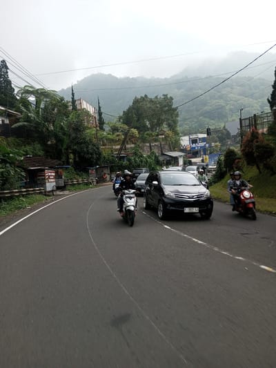 The image shows a winding road with several motorbikes and a black car traveling along it. The location appears to be a mountainous area, possibly in Mae Chan District, Thailand. The weather is overcast with low-hanging clouds obscuring the mountain peaks. Lush greenery and trees line the roadside. Buildings and small shops are visible along the left side of the road. Traffic appears moderate. The car in the foreground has a license plate number "F 1821 LJ." The motorbikes have riders, most wearing helmets.