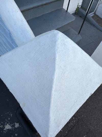 An exterior shot captures a white, pyramid-shaped concrete cap atop a pillar, with a section of grey outdoor stairs and railing visible in the background. The scene is likely located in Torquay, United Kingdom. The concrete has a rough texture with some minor staining and discoloration. The stairs are worn and appear to be made of concrete, with a dark grey railing running alongside them. A section of white brick wall and a window are also visible behind the stairs. The ground below the pillar is dark asphalt. No people or animals are present in the image, and no actions are depicted. The overall impression is one of an outdoor architectural feature in a residential or public area.