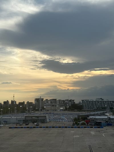 An elevated, forensic, matter-of-fact description of a cityscape in Pak Kret City Municipality, Thailand, captured from a high vantage point. The scene is dominated by a vast expanse of concrete in the foreground, marked with white lines indicating parking bays and tire tracks, suggesting a commercial or recreational area. Beyond the concrete, a large outdoor karting track is visible, delineated by blue and white barriers. Several buildings are clustered in the middle ground, including a structure labeled "IMPACT SPEED PARK," indicating a leisure facility. The background features a dense urban skyline with a variety of buildings, ranging from modern high-rises to older structures. Tall communication towers pierce the skyline, and a large, multi-story building with a distinctive layered facade stands prominently. The sky above is dramatic, filled with heavy, dark clouds interspersed with patches of bright, warm light, suggesting either the aftermath of a storm or a sunset with significant cloud cover. A faint contrail from an aircraft is visible high in the sky. No people are clearly discernible in the image, and no specific actions or interactions are observable. The overall environment appears to be an outdoor public space within a developed urban area.